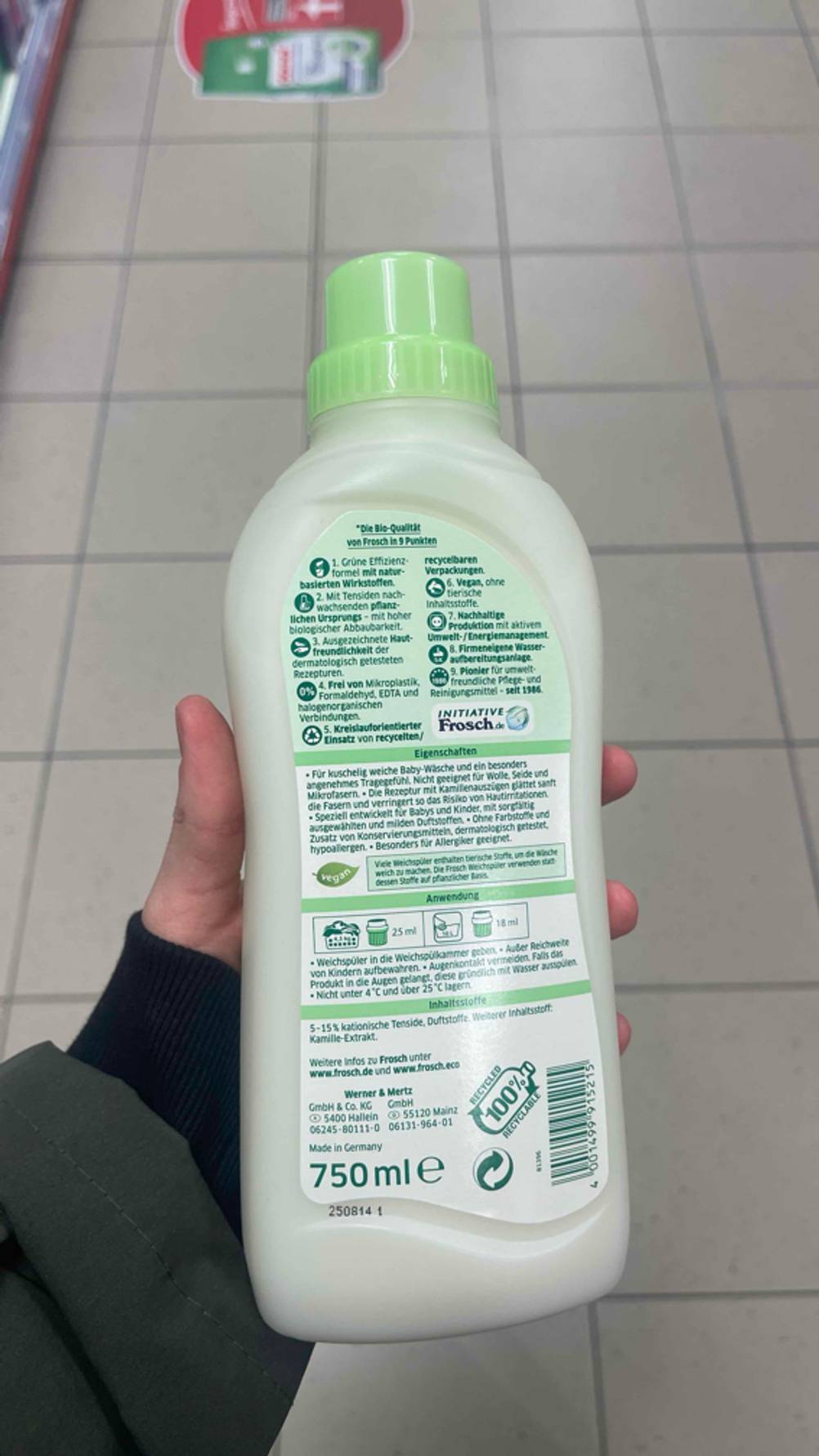 Eine Hand hält eine weiße Flasche inklusive der Produktbeschreibung des getesteten veganen Weichspülers über einem weißen Fliesenboden.