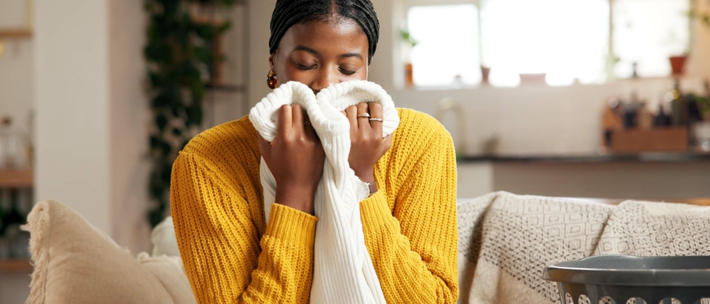 Eine entspannte Frau zu Hause, umgeben von frisch gewaschener Wäsche. Sie genießt den frischen Duft von Weichspüler, während sie im komfortablen Wohnzimmer sitzt. Die Atmosphäre ist geprägt von Frische, Sauberkeit und einem Gefühl von Wohlbefinden.