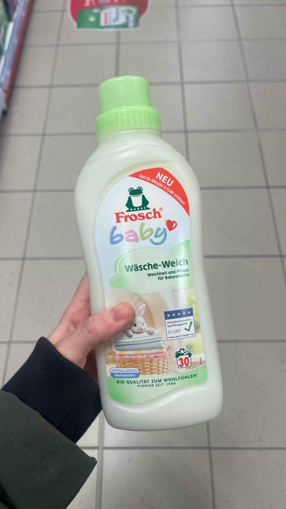 Eine Hand hält eine weiße Flasche mit dem getesteten veganen Weichspüler über einem weißen Fliesenboden.