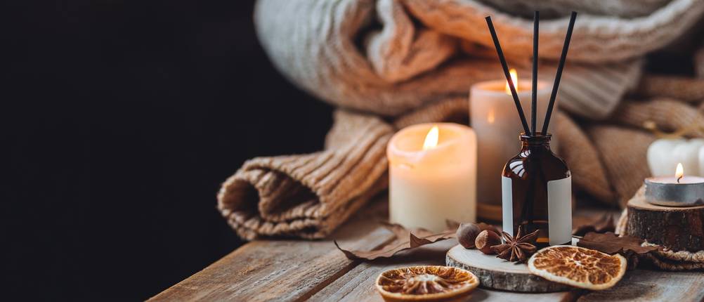 Auf einem Holztisch liegen getrocknete Orangenscheiben, ein Fläschchen mit ätherischem Öl und Holzstäbchen, Kerzen und einer Wolldecke.