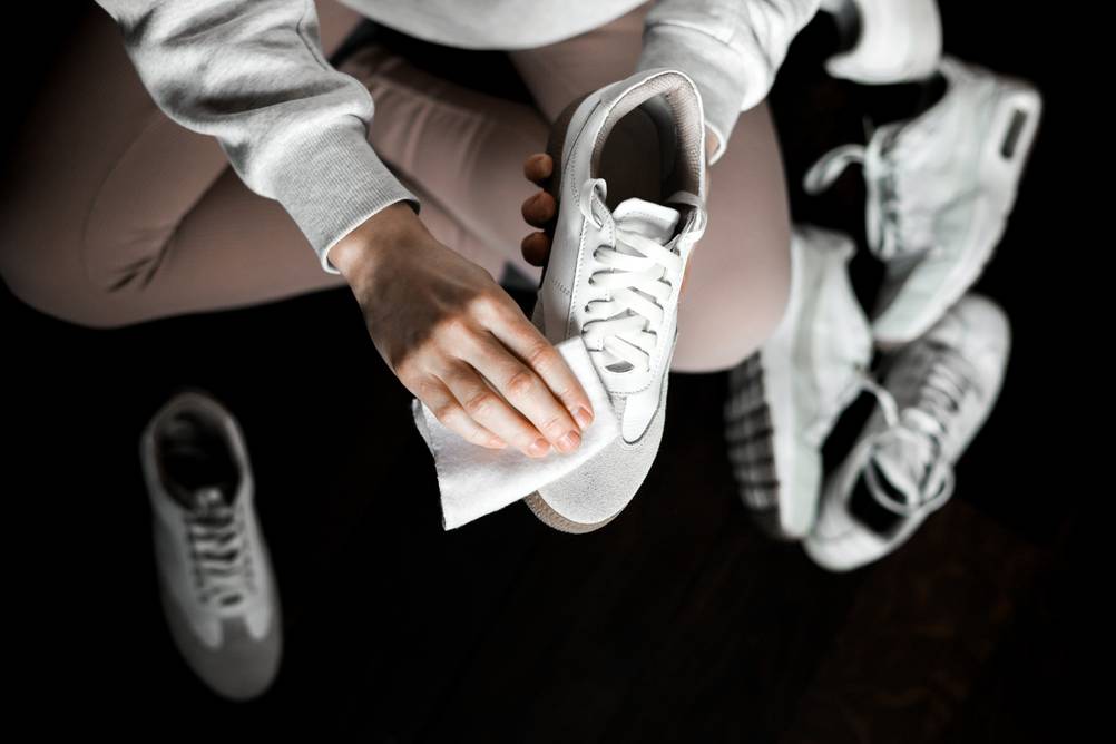 Eine Frau sitzt im Schneidersitz auf dem Boden und reinigt mit einem Tuch einen weißen Sneaker. Um sie herum verstreut liegen weitere weiße Sneaker und Turnschuhe