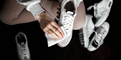 Eine Frau sitzt im Schneidersitz auf dem Boden und reinigt mit einem Tuch einen weißen Sneaker. Um sie herum verstreut liegen weitere weiße Sneaker und Turnschuhe