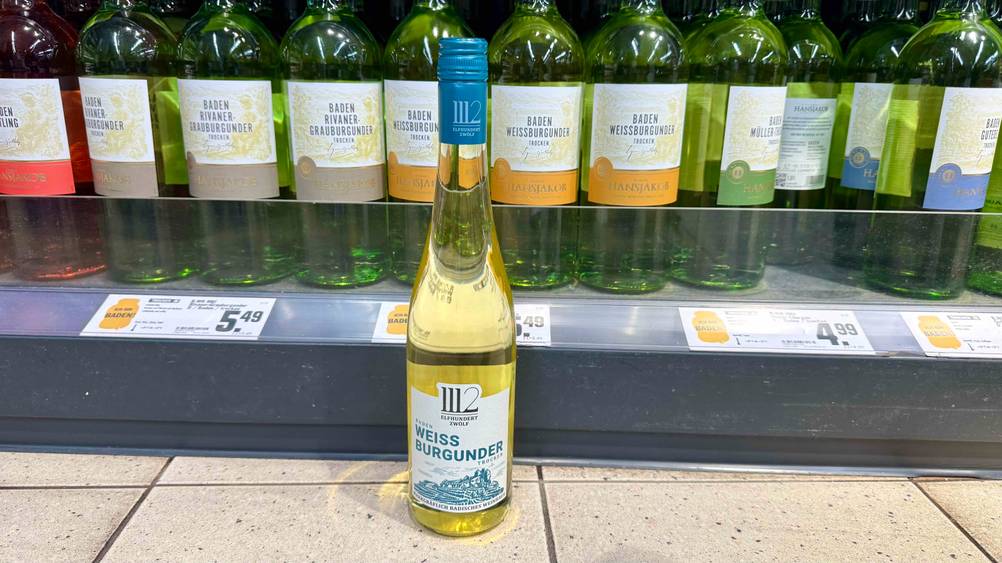 Weißwein (trocken) im Test: Eine Flasche vor eine Verkaufsregal mit Preisangaben auf dem Boden stehend.
