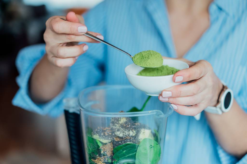 Weizengraspulver-Test: Eine Frau gibt mit einem Löffel Weizengraspulver in einen Smoothie-Maker. 