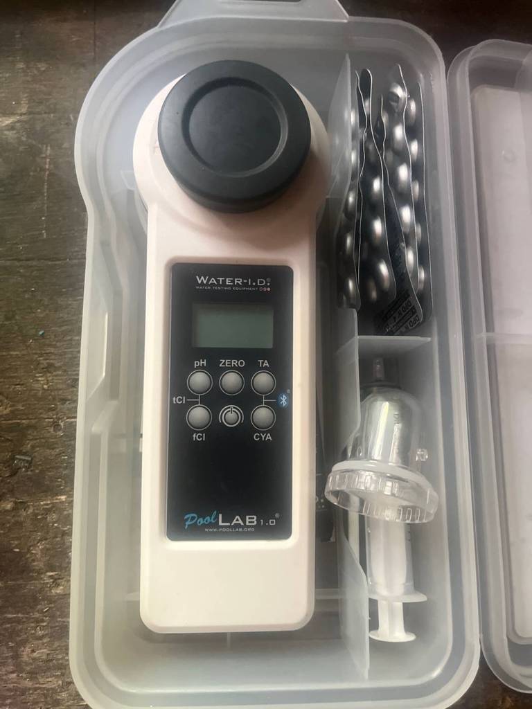 Ein getesteter Pooltester des Typs PoolLab 1.0, mit Blick auf die Anzeige und die Bedientasten, liegt inklusive Zubehör in einer transparenten Aufbewahrungsbox auf Holz.