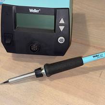 Der Weller WE 1010 Lötkolben liegt mit angeschlossener Station auf einer Holzoberfläche. Die digitale Bedieneinheit und der ergonomische Griff des Lötkolbens sind deutlich erkennbar.