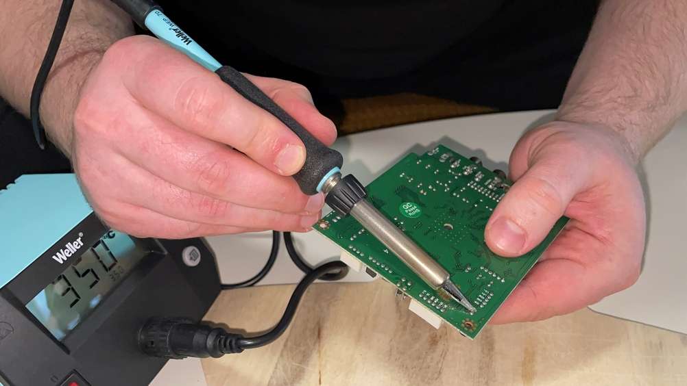 Ein Nutzer lötet mit dem Weller WE 1010 eine grüne Leiterplatte, während das Steuergerät im Hintergrund eine Temperatur von 350 °C anzeigt. Die Hände führen den Lötkolben präzise an eine feine Lötstelle.