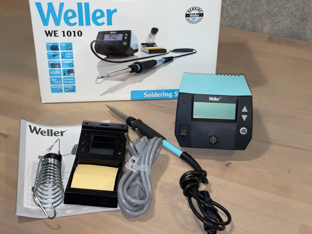 Die komplette Ausstattung des Weller WE 1010 ist ausgebreitet: Steuergerät, Lötkolben, Ablage mit Reinigungsschwamm, Netzanschluss, Bedienungsanleitung und Originalverpackung. Alle Komponenten wirken hochwertig verarbeitet und sofort einsatzbereit.