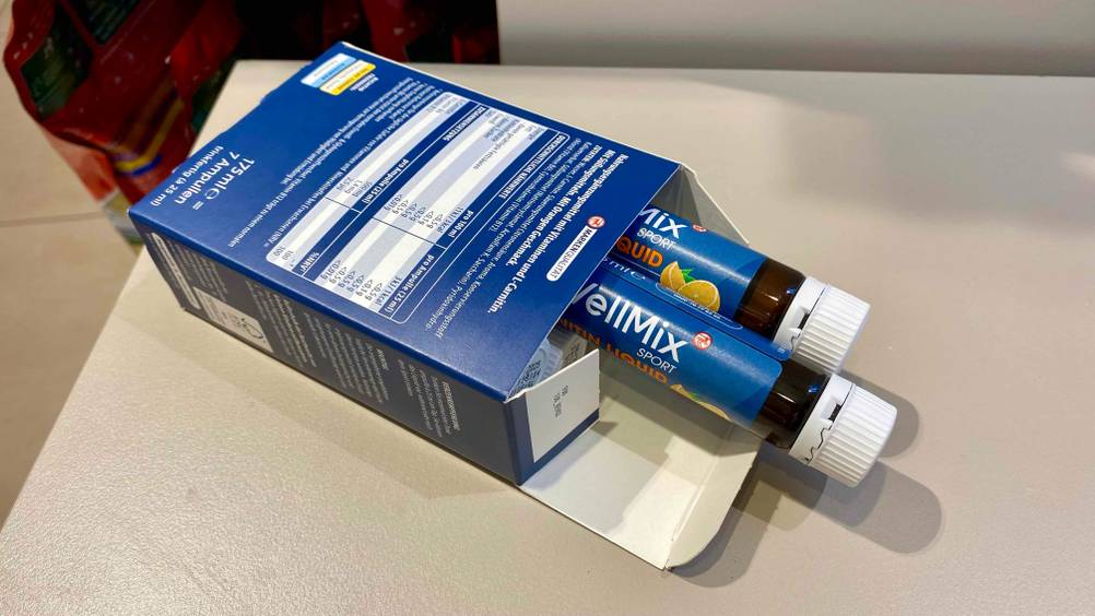 Im Test: Zwei Röllchen L-Carnitin ragen aus der Originalverpackung, die mit der Rückseite nach oben auf einer weißen Fläche liegt.