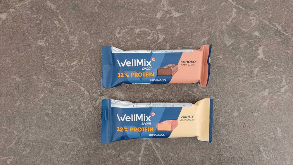 Zwei Proteinriegel im Test in unterschiedlichen Sorten liegen nebeneinander auf einer grauen Fläche.