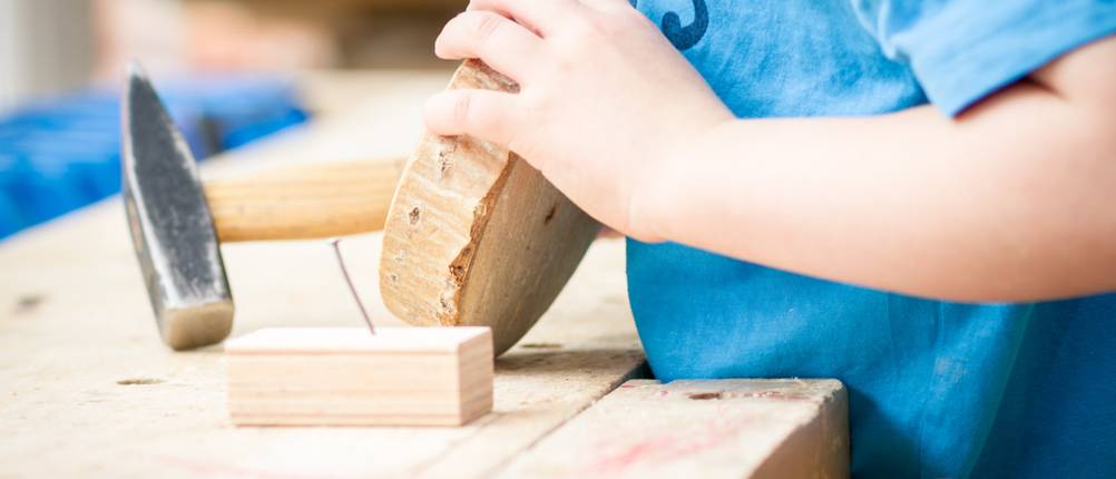 Kinder-Werkbank im Test: Hier ein Modell aus Massivholz, mit einem Holzstück welches von einem Kind mit Hammer bearbeitet wird. 