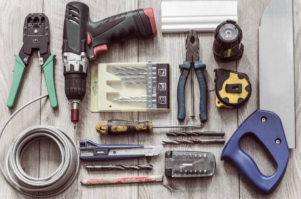 Werkzeugkoffer mit Akkuschrauber im Test: Einige Werkzeuge, wie Zangen, Säge, Cutter, Schraubendreher sowie ein Kabel, ein Stückfußleiste und ein Maßband auf einem Holzuntergrund.