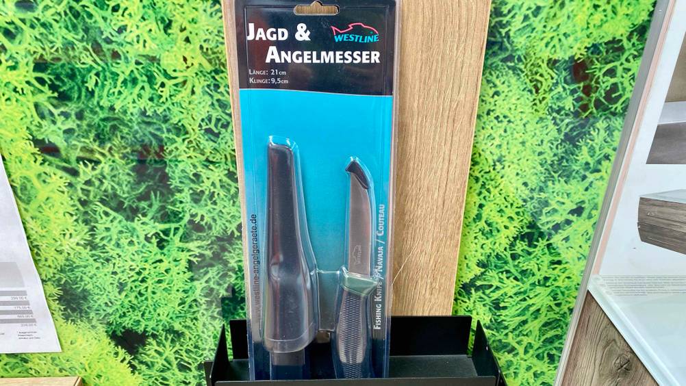 Getestet: Ein verpacktes Jagdmesser steht in einer schwarze Schale vor einem Plakat.