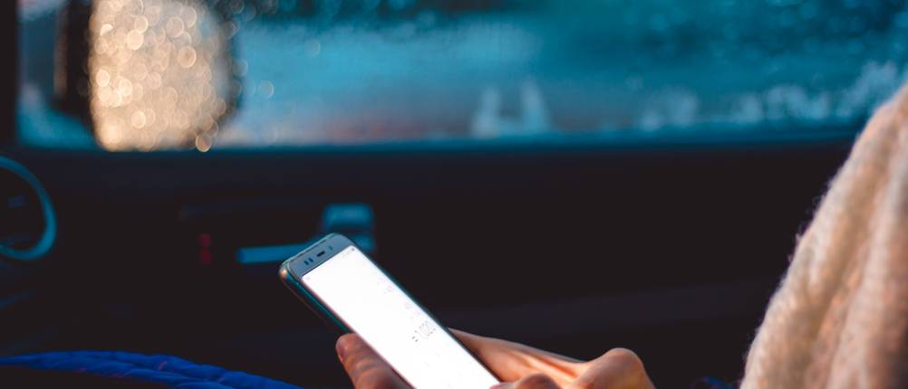 Person sitzt im parkenden Auto bei Regen und schaut auf helles Handydisplay.