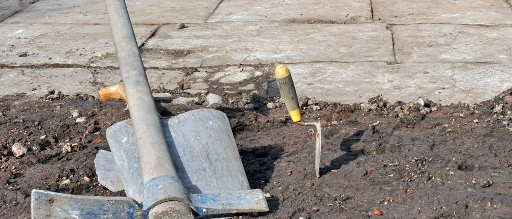 Wiedehopfhacke im Test: Eine Wiedehopfhacke, eine Kelle und eine Schaufel liegen auf nackter Erde; dahinter eine Fläche mit Stein oder Betonplatten.
