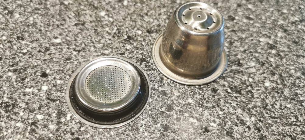Eine getestete wiederverwendbare Kaffeekapsel auf dem Kopf stehend neben der Verschlusskappe mit Löchern.