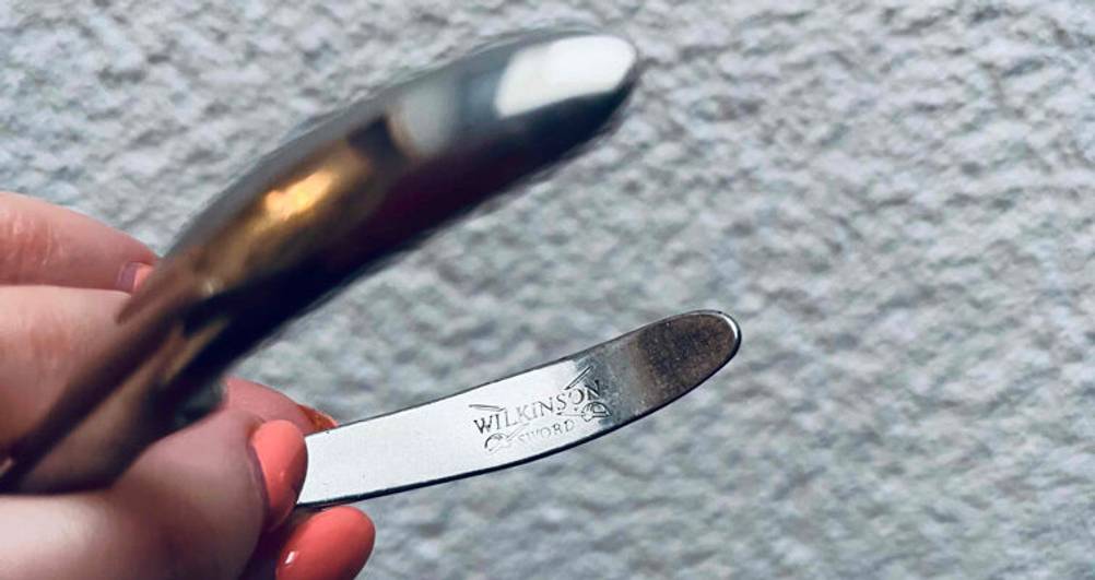 Eine Frauenhand hält das Wilkinson Markenzeichen auf einer Edelstahl-Nagelhautzange im Test vor einer weißen Rauputzwand in die Kamera.