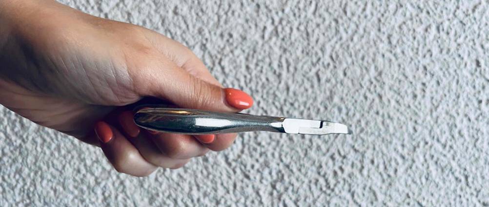 Eine Frauenhand hält eine geschlossene Edelstahl-Nagelhautzange im Test vor eine weiße Rauputzwand.