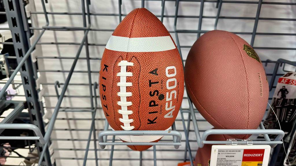 Der getestete Football in Rot mit weißer Naht liegt neben einem einfarbigen Football im Verkaufsregal.