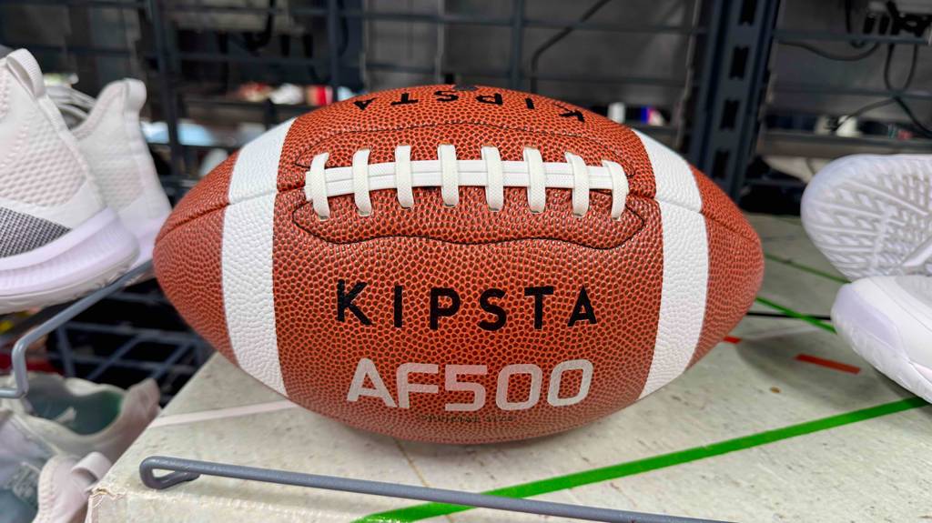 Football-Bälle im Test: Ein rotes Modell AF500 von KIPSTA liegt auf einem Verkaufsregal zwischen weißen Sportschuhen.