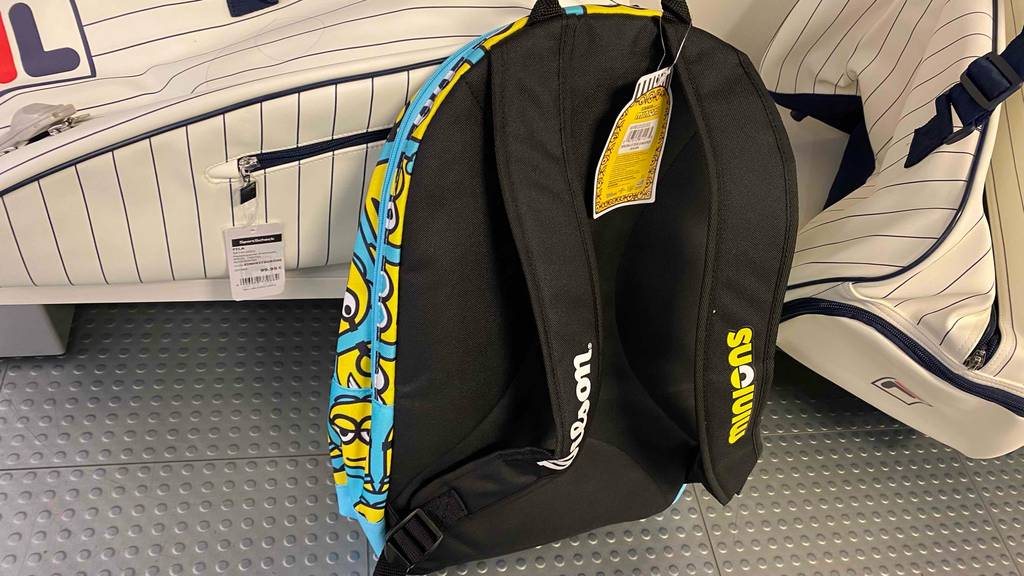 Kinder-Tennisrucksack im Test: Ein hellblauer Rucksack mit gelben Minion-Figuren lehnt an einem Verkaufsregal mit weiß-schwarz gestreiften Tennistaschen. Im Vordergrund sind die schwarzen gepolsterten Tragegurte.