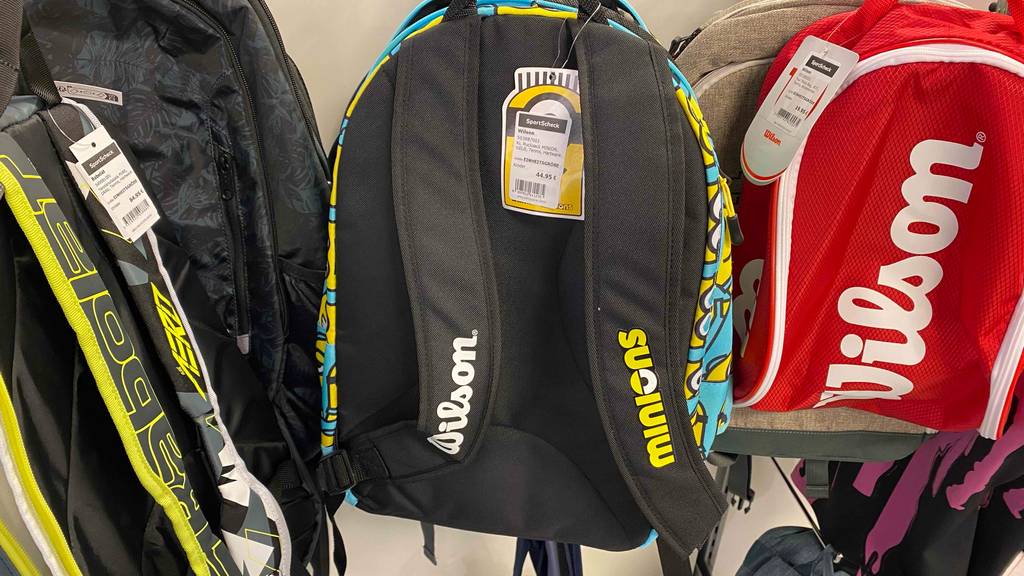 Tennisrucksack-Kinder-Test: An einem Verkaufsstand hängen unterschiedliche Rucksäcke, mit Minions-Motiv, Wilson-Motiv und weitere, mit Fokus auf gepolsterten Schultergurten.