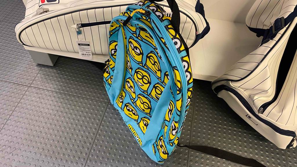 Tennisrucksack für Kinder im Test: Ein hellblauer Rucksack mit gelben Mignon-Figuren lehnt an einem Verkaufsregal mit weiß-schwarz gestreiften Tennistaschen.