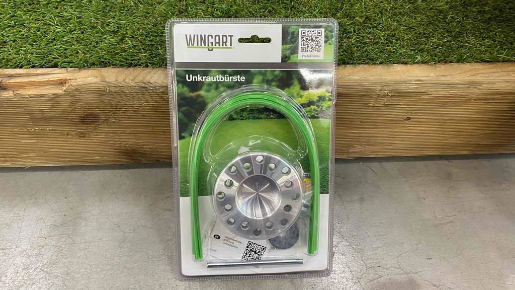 Im Test: verpackte Wingart Unkrautbürste lehnt an Holzlatte.