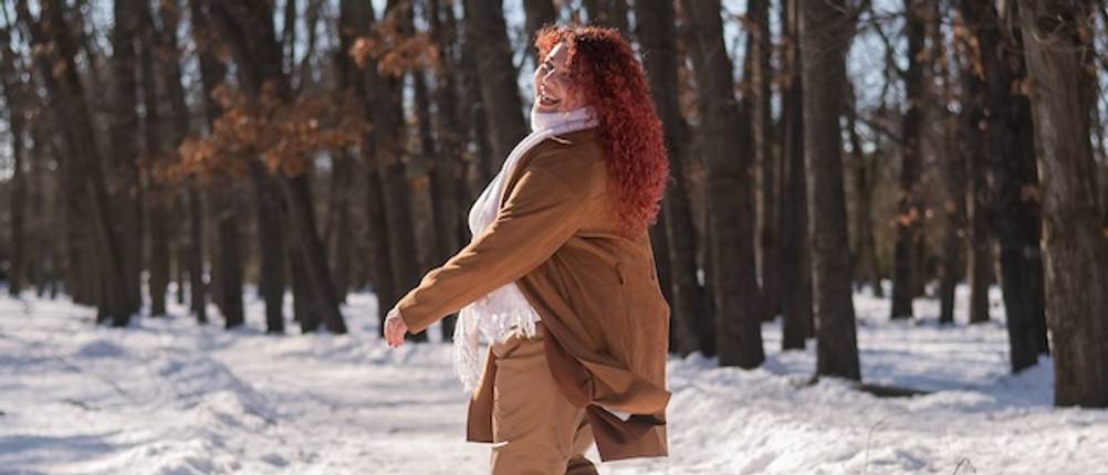 Warme Damen-Winterjacke im Test: Eine fröhliche Frau mit roten Haaren trägt eine Winterjacke in Braun in einer Schneelandschaft.