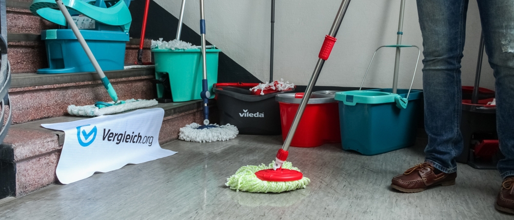 Eine Person wischt den Boden mit einem Wischmopp vor einer Treppe, im Hintergrund stehen weitere Eimer mit Mopp.