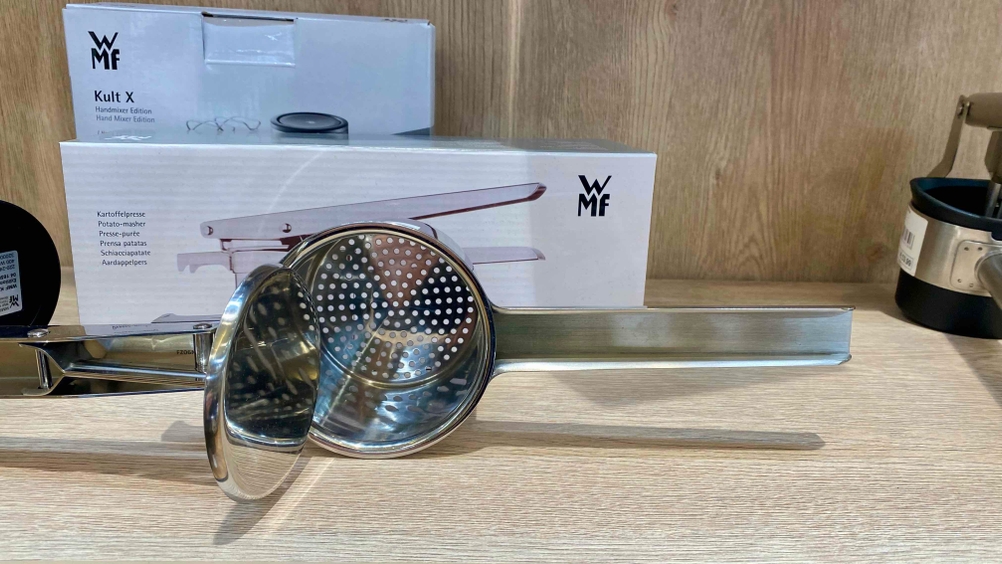 Eine auf der Seite liegende geöffnete Kartoffelpresse im Test mit Fokus das Sieb, auf einer Holzfläche, im Hintergrund Originalkartons.
