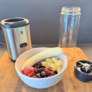 Alle Teile des Smoothie-Makers stehen gemeinsam mit einer Schüssel Obst auf einem Holzbrett.