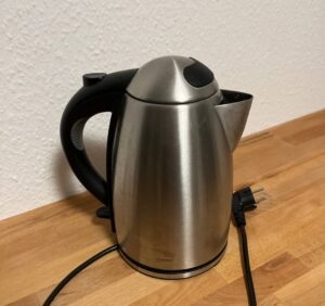 Ein Wasserkocher aus Edelstahl mit schwarzem Kunststoffgriff steht inklusive Kabel auf einer Holzfläche.