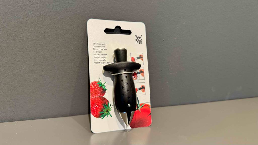 Ein getesteter Tomatenstrunkentferner in Schwarz lehnt inklusive Packpappe an einer grauen Wand.