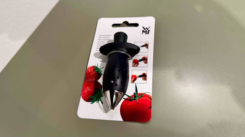 Tomatenstrunkentferner-Test: Ein schwarzes Modell der Marke WMF liegt oberhalb der Verpackung auf einem grauen Regalboden.