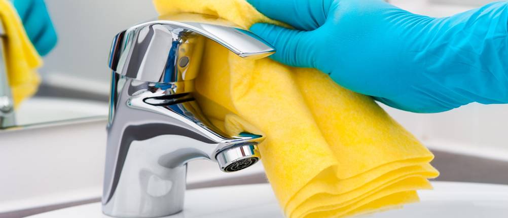 Behandschuhte Hand reinigt den Wasserhahn mit einem Putzlappen.