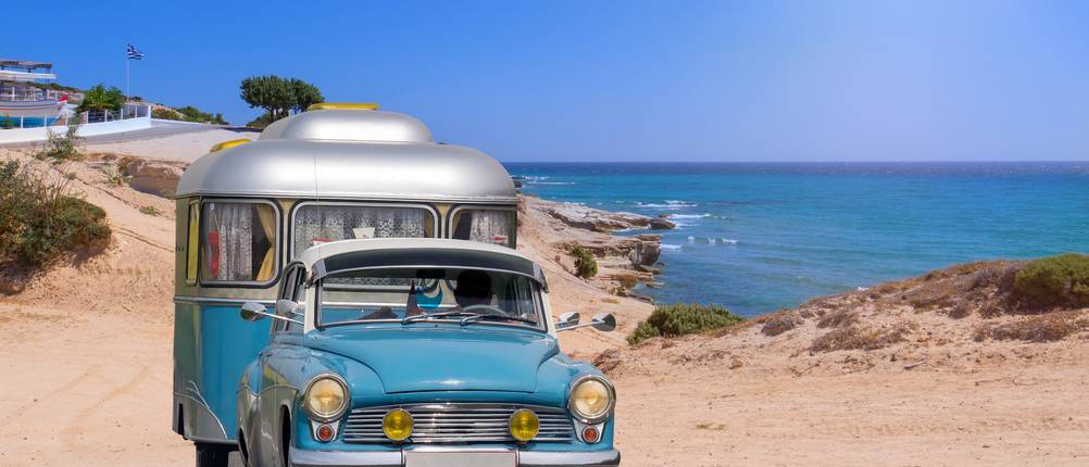 Ein Oldtimer mit einem Wohnwagen fährt an einem Strand entlang.