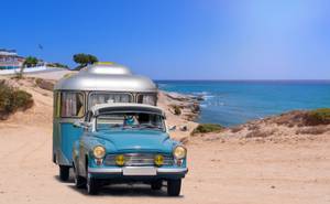 Wohnwagenversicherungen im Test: Ein Oldtimer mit einem Wohnwagen fährt an einem Strand entlang.
