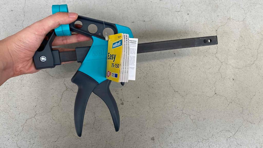 Im Test: Hand hält eine Wolfcraft Einhandzwinge vor grauer Wand.