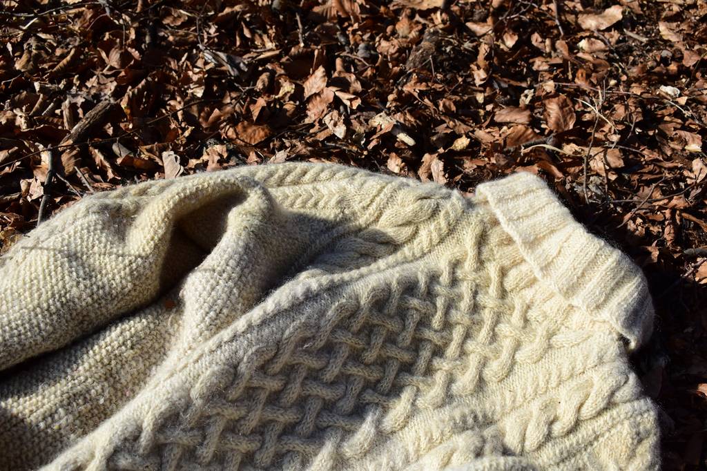 Weißer Wollpullover mit Zopfmuster liegt auf braunem Herbstlaub.
