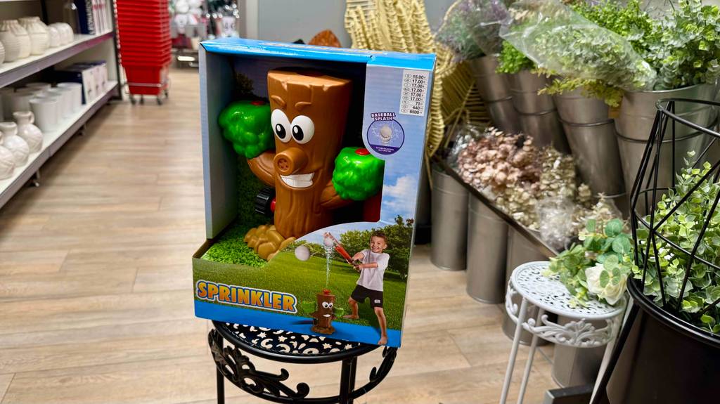 Der getestete Wassersprinkler für Kinder im Baumstamm-Design steht innerhalb einer Verpackung auf einem Blumentisch im Gartencenter.