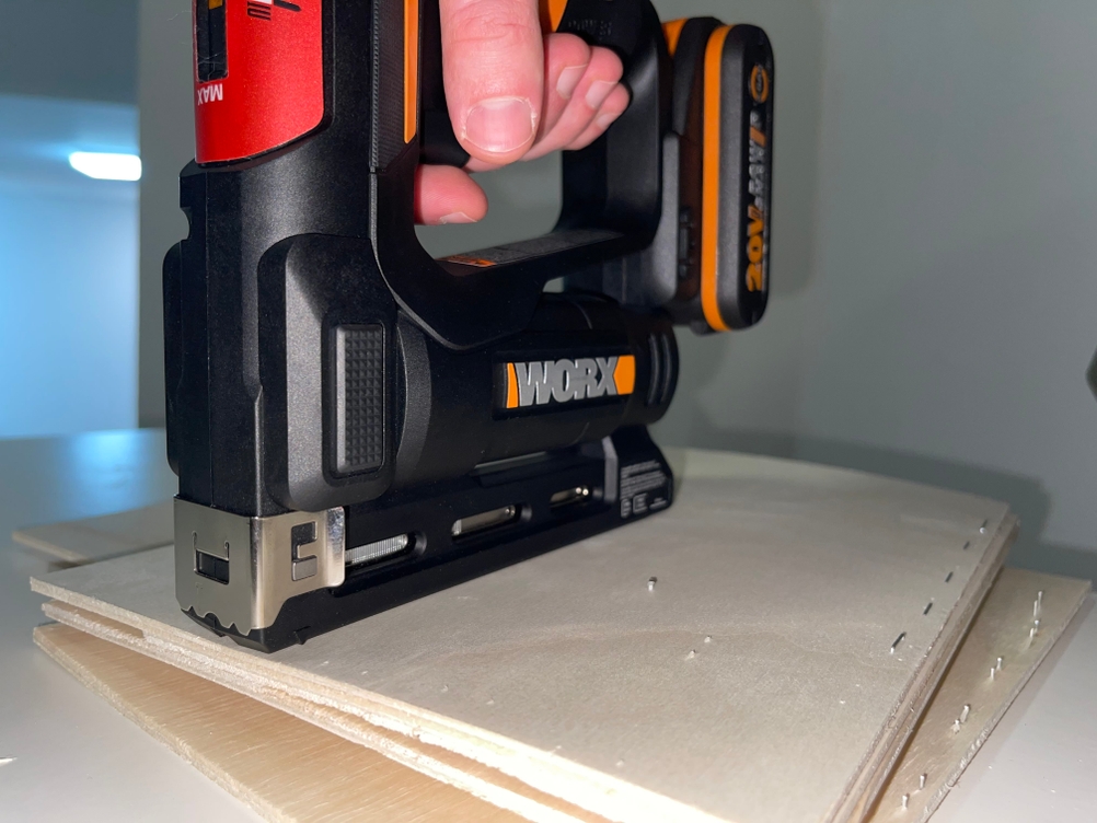 Seitliche Ansicht des WORX WX843.9 Akku-Tackers bei der Anwendung auf zwei Holzplatten. Die Hand führt das Gerät beim Tackern, Klammern sind bereits eingeschossen.