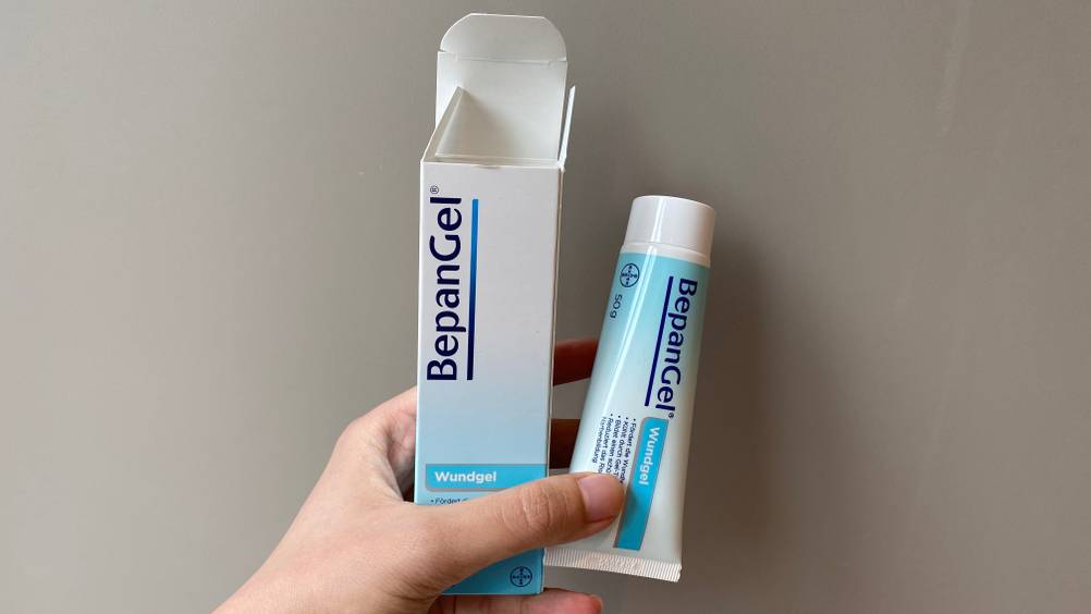 Eine Hand hält eine Tube und deren Verpackung des getesteten Wundgels vor eine graue Wand.