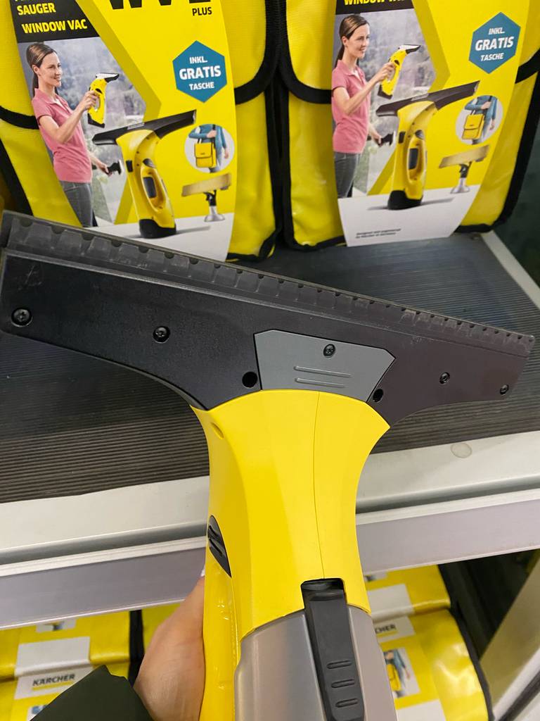 Hand hält gelben, getesteten Kärcher-Fenstersauger, mit Fokus auf die Saugeinheit, vor ein Verkaufsregal mit original verpackten Kärcher-Fenstersaugern.