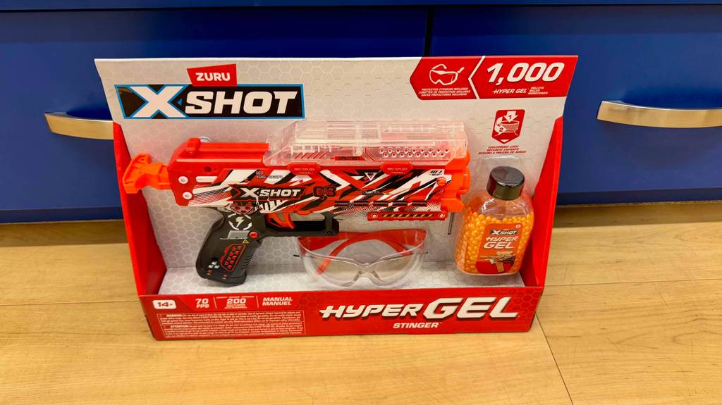 Gel-Ball-Blaster-Test: Das Set, bestehend aus einer Pistole, einer Sicherheitsbrille und einer Dose mit Gel-Kugeln, befindet sich in seiner Verpackung auf dem Boden vor einem blauen Verkaufsregal.