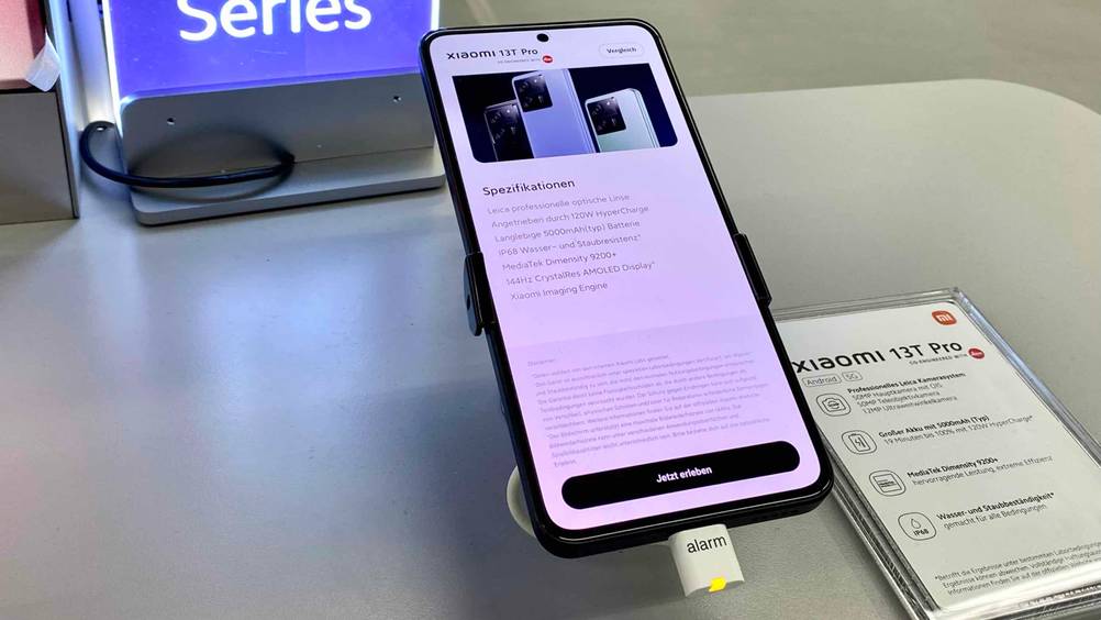 Nahaufnahme eines getesteten Xiaomi-Handys in einer Verkaufsfläche mit angeschaltenem Display und einer Infotafel daneben.