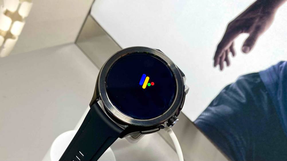 Nahaufnahme des Displays einer getesteten Xiaomi-Smartwatch vor einem Plakat mit einer Hand.