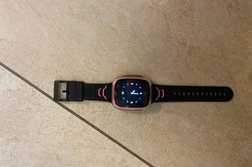 Eine eingeschaltete, getestete Kinder-Smartwatch in Schwarz inklusive Armband liegt auf einem hellen Fliesenboden.