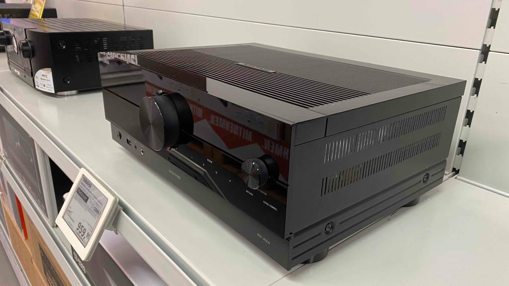 Schräge Seitenansicht auf einen schwarzen Yamaha-Receiver im Test, mit Fokus auf den Einstellregler und das Preisschild, steht neben einem anderen Modell auf einem hellen Verkaufsregal.