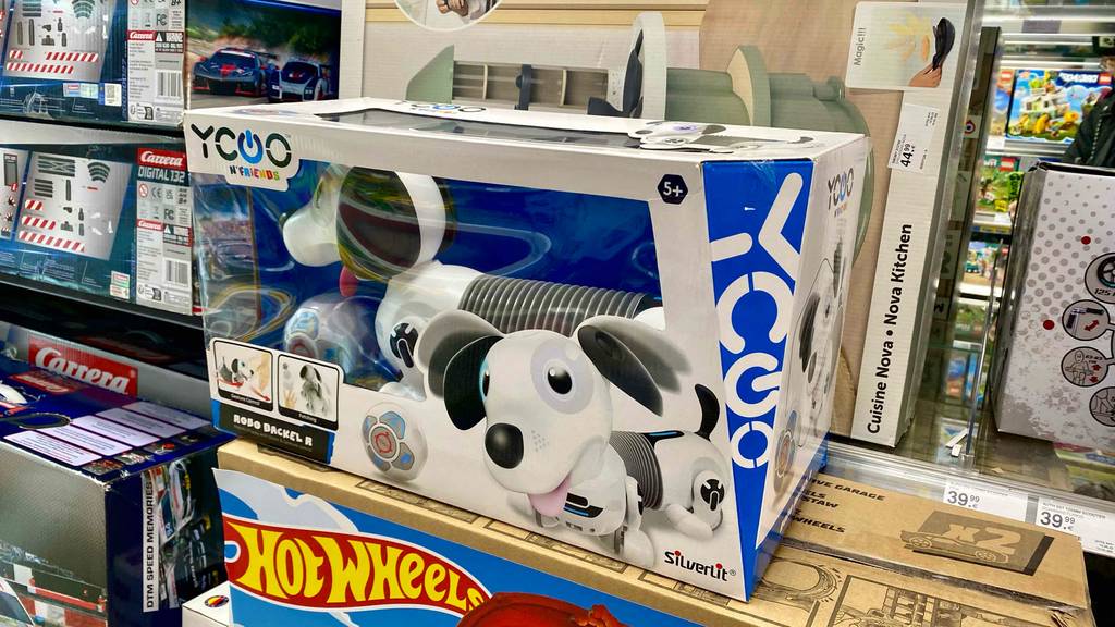 In einem Geschäft steht vor anderen technischen Spielen ein Roboterhund im Test: der Yoco Robo Dackel in weiß-schwarzem Design ist in seinem Karton mit Sichtfenster.